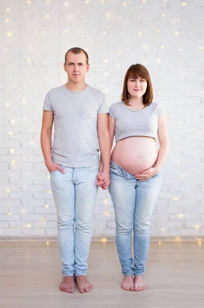 Ganzkörperporträt Von Schwangerem Paar Posiert Über Weißer Ziegelwand Mit Weihnachtsbeleuchtung — Stockfoto