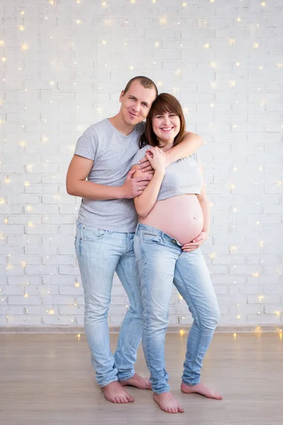 家庭与亲子关系概念 怀孕夫妇的全长肖像 挂在白色砖墙上 有圣诞彩灯 — 图库照片