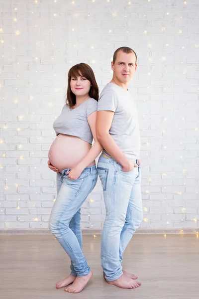 親の概念 クリスマスライトと白いレンガの壁にかわいい妊娠中のカップルの完全な長さの肖像画 — ストック写真
