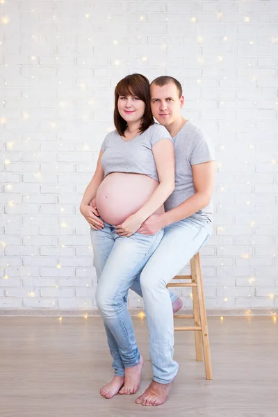 家庭概念 可爱的怀孕夫妇在白色砖墙上的肖像 圣诞彩灯 — 图库照片