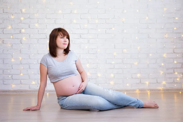 家庭和母性概念 孕妇坐在白色砖墙的地板上 圣诞灯火通明 — 图库照片