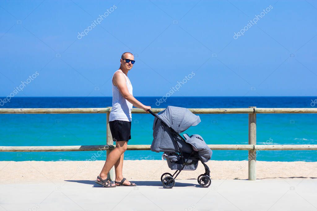 parenting and travel concept - side view of young man pushing baby stroller on the summer beach