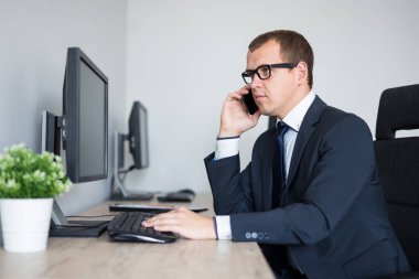 Modern ofiste bilgisayar kullanan ve telefonla konuşan yakışıklı iş adamının portresi.