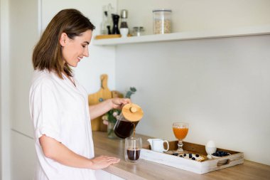 Kahvaltı konsepti: Pijamalı bir kadın modern mutfakta kahve makinesinden kahve dolduruyor.