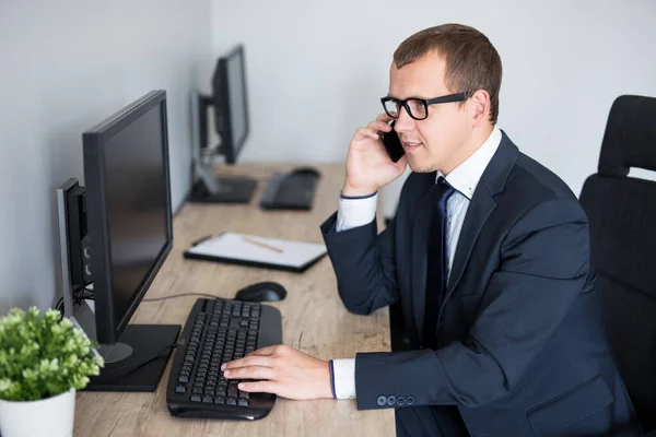 コンピュータを使って現代のオフィスで電話で話すハンサムなビジネスマン — ストック写真