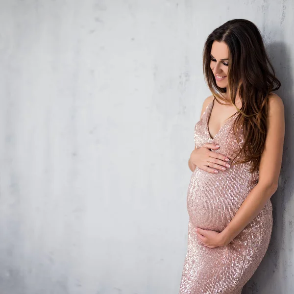 幸せな可愛いです妊娠中の女性で光沢のある夜服見ます彼女のおなか上の灰色の壁の背景とともにコピースペース — ストック写真