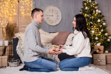 Noel, yeni yıl ve aşk konsepti. Yakışıklı bir adam Noel ağacı, hediye kutuları ve çelenk ışıklarıyla süslü odada kız arkadaşına Noel hediyesi veriyor.