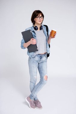 Dişleri, dizüstü bilgisayarı, kahve fincanı ve gri arka planda sırt çantası olan gülümseyen kadın öğrenci.