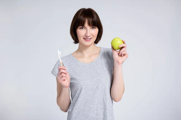 歯と歯列矯正の概念 歯の上にブレースを持つ若い女性の肖像画灰色の背景の上に緑のリンゴと歯ブラシを保持 — ストック写真