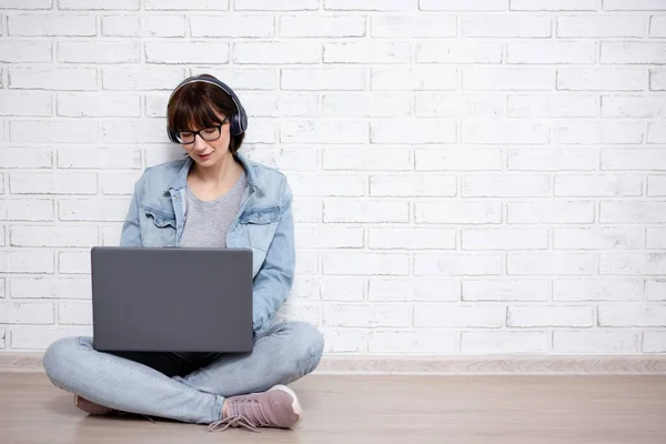 若い女性や10代の少女が床に座ってノートパソコンを使いコピースペースのある白いレンガの壁の背景の上にヘッドフォンで音楽を聴く — ストック写真