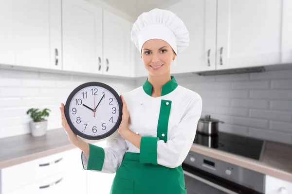 时间概念 现代厨房里穿着厨师制服的年轻貌美的女人和办公室钟表 — 图库照片