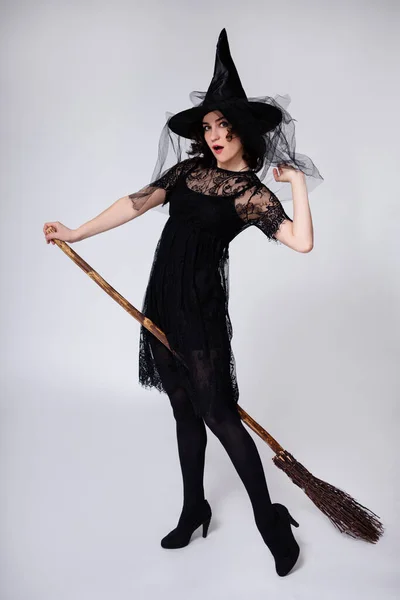 黒魔女ハロウィンの衣装で驚きの美しい女性の完全な長さの肖像画と帽子とともに座ってほうき上の白い背景 — ストック写真