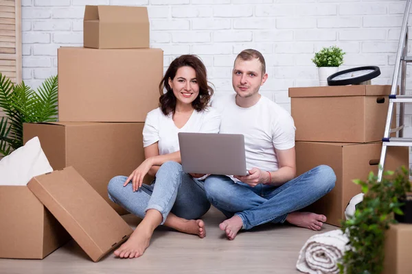 moving day concept - happy young couple with laptop and moving boxes in their new house or flat