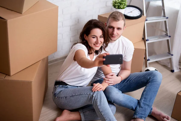 moving day - portrait of young smiling couple taking selfie photo with smartphone, surrounded cardboard boxes in new house or flat