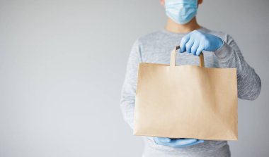 Coronavirus ve temas kurulamayan teslimatçı - tıbbi maskeli genç teslimatçı ve beyaz arka planda fotokopi alanı olan kağıt torba tutan eldivenler