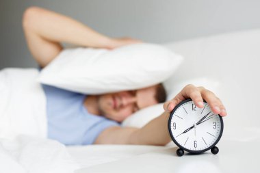 Akşamdan kalmalık ve rutin konsept. Bir adam alarmlı saatle uyanıyor ve yatak odasında yastıkla başını örtüyor.