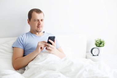 Evdeki yatakta akıllı telefon kullanan mutlu yakışıklı adamın portresi.