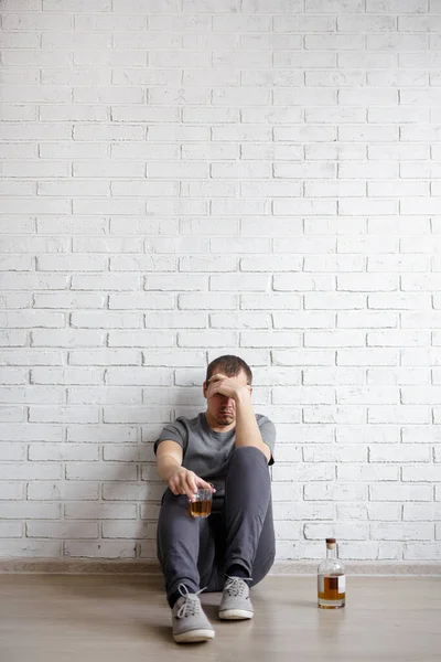 アルコール依存症の概念 コピースペースと白いレンガの壁の背景の上に床に座っているウイスキーのボトルとガラスを持つストレスのある男 — ストック写真