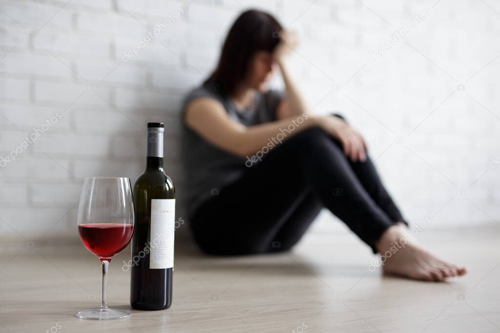female alcoholism, relationship, divorce and depression concept - close up of glass and bottle of wine and stressed woman crying and sitting on the floor at home
