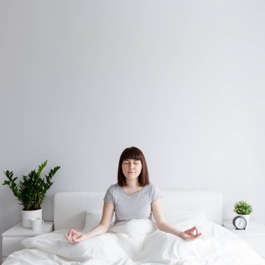 Evde yoga pozisyonunda yatakta oturan mutlu bir kadın, beyaz duvar arkaplanının üzerinde boşluğu kopyalayarak
