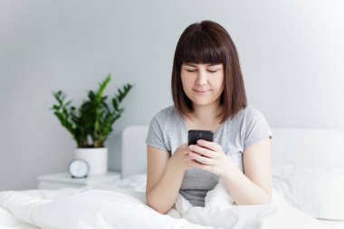 Mutlu bir kadının portresi yatakta oturuyor ve evde akıllı telefon kullanıyor.
