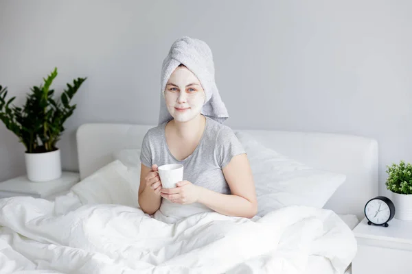 朝とスキンケアのコンセプト 顔のマスクをした若い女性がベッドに座ってコーヒーを飲んで — ストック写真
