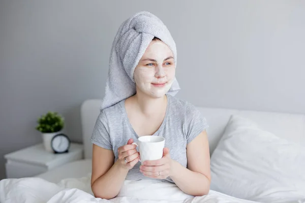 朝とスキンケアのコンセプト 顔のマスクとコーヒーカップとベッドに座っている若い女性の肖像画 — ストック写真