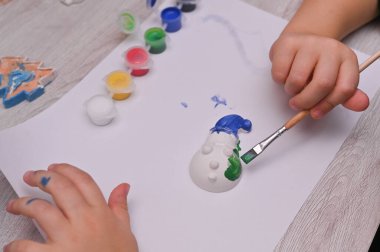 Çocuk, alçıtaşı oyuncakları üzerinde parlak renklerde fırça ile basit fırça darbeleri kullanır. Kız alçıdan kardan adam figürü çiziyor. DIY Noel Süslemeleri.