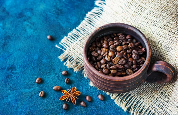 Una taza de barro llena de granos de café de pie sobre la mesa, con estrellas de anís y una cuchara de madera llena de café molido —  Fotos de Stock