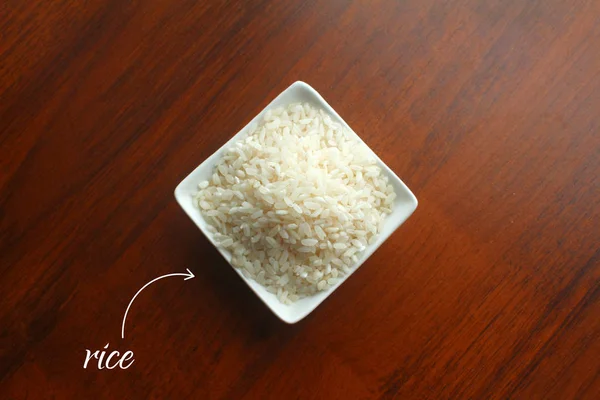 Taça branca com grãos de arroz branco em uma mesa de madeira, vista superior — Fotografia de Stock