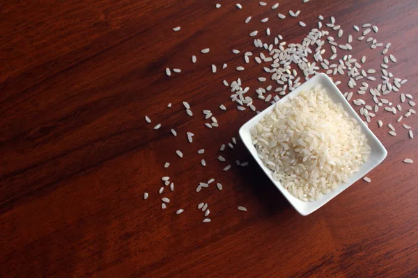 Taça branca com grãos de arroz branco em uma mesa de madeira, vista superior. Grãos espalhados aleatoriamente ao redor da tigela . — Fotografia de Stock
