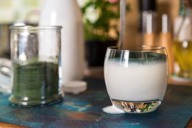 Closeup açık cam ve yeşil spirulina tozu mutfak masasının üzerinde karıştırmak hazır kaşıkla taze kefir probiotik içki görüntülemek
