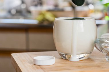 Closeup açık cam ve yeşil spirulina tozu mutfak masasının üzerinde karıştırmak hazır kaşıkla taze kefir probiotik içki görüntülemek