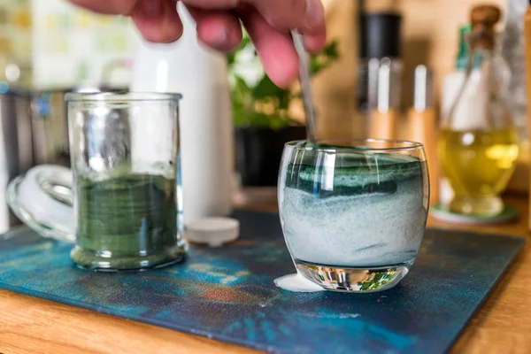 Вид крупным планом свежий кефир пробиотик напиток в прозрачном стекле и ложка с зеленой спирулиной порошок готов смешать на кухонном столе — стоковое фото