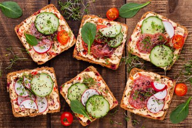 Krem peynirli, sebzeli ve salamlı sandviç. Salatalıklı sandviçler, turp, domates, gri arka planda salam, üst manzara. Düz yatıyordu.