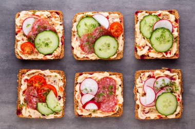 Krem peynirli, sebzeli ve salamlı sandviç. Salatalıklı sandviçler, turp, domates, gri arka planda salam, üst manzara. Düz yatıyordu.