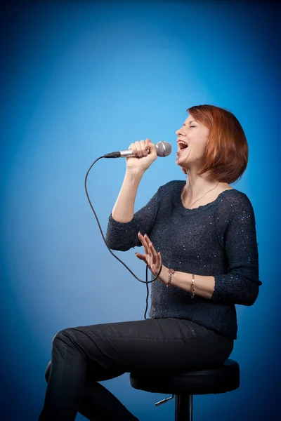 red-haired woman in black clothes with a microphone sits on a bar stool and sings on a blue background