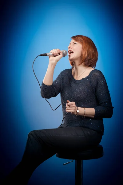 red-haired woman in black clothes with a microphone sits on a bar stool and sings on a blue background