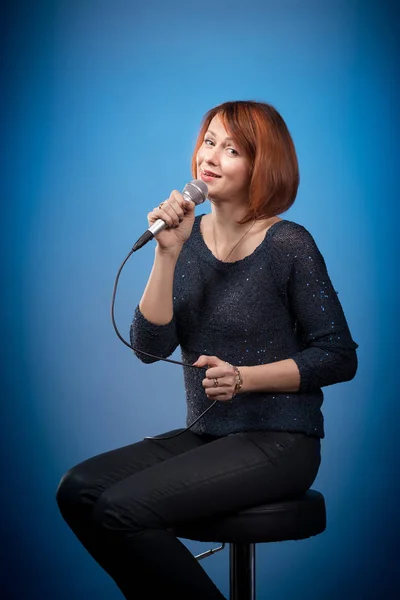 red-haired woman in black clothes with a microphone sits on a bar stool and sings on a blue background