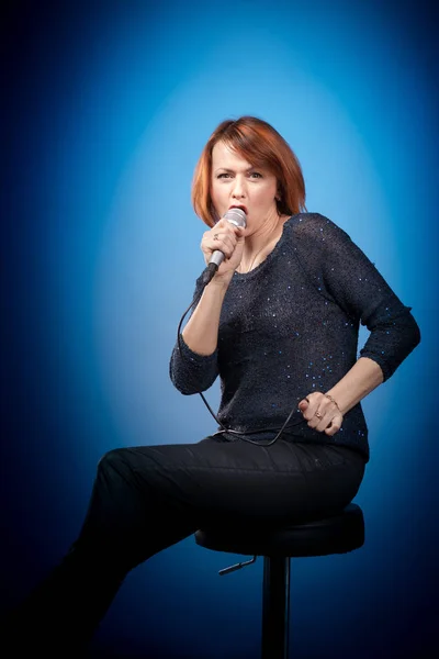 red-haired woman in black clothes with a microphone sits on a bar stool and sings on a blue background