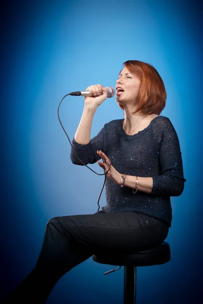 red-haired woman in black clothes with a microphone sits on a bar stool and sings on a blue background