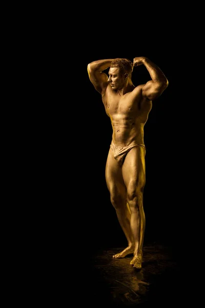 Male athlete bodybuilder in golden bodyart posing on black background