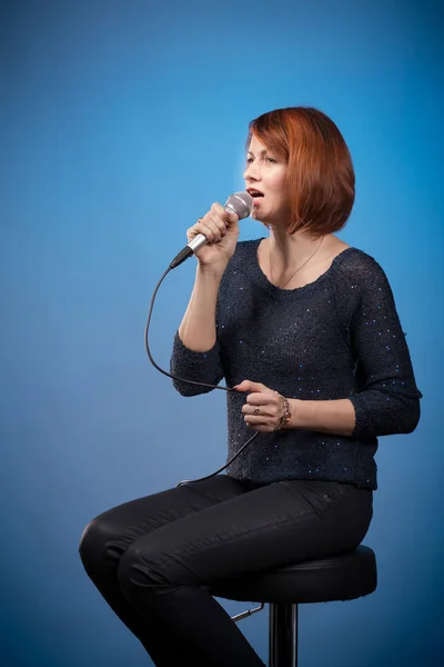 red-haired woman in black clothes with a microphone sits on a bar stool and sings on a blue background