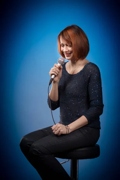 red-haired woman in black clothes with a microphone sits on a bar stool and sings on a blue background