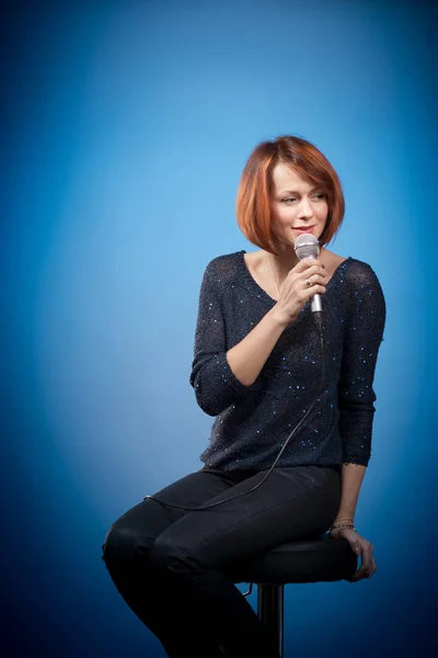 red-haired woman in black clothes with a microphone sits on a bar stool and sings on a blue background