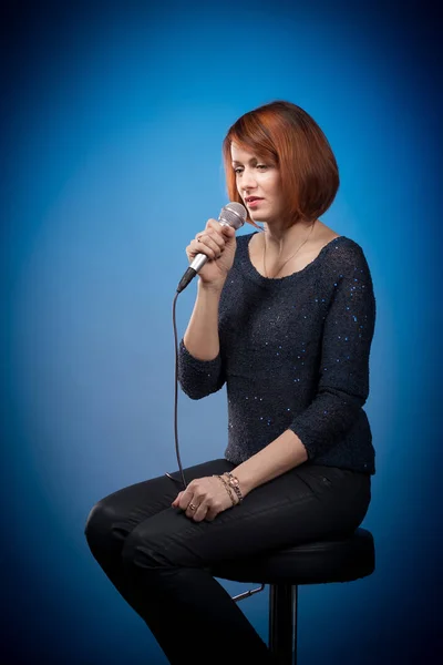 red-haired woman in black clothes with a microphone sits on a bar stool and sings on a blue background