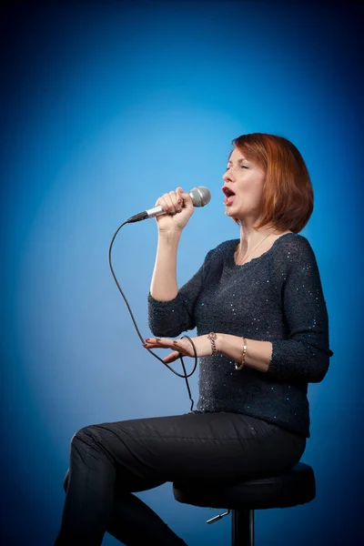 red-haired woman in black clothes with a microphone sits on a bar stool and sings on a blue background