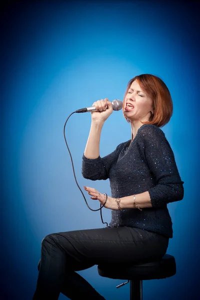 red-haired woman in black clothes with a microphone sits on a bar stool and sings on a blue background