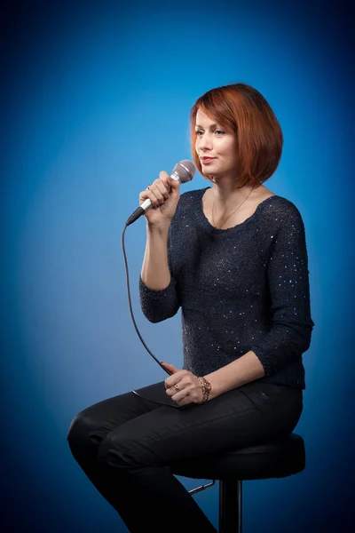 red-haired woman in black clothes with a microphone sits on a bar stool and sings on a blue background