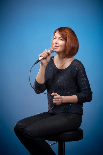 red-haired woman in black clothes with a microphone sits on a bar stool and sings on a blue background